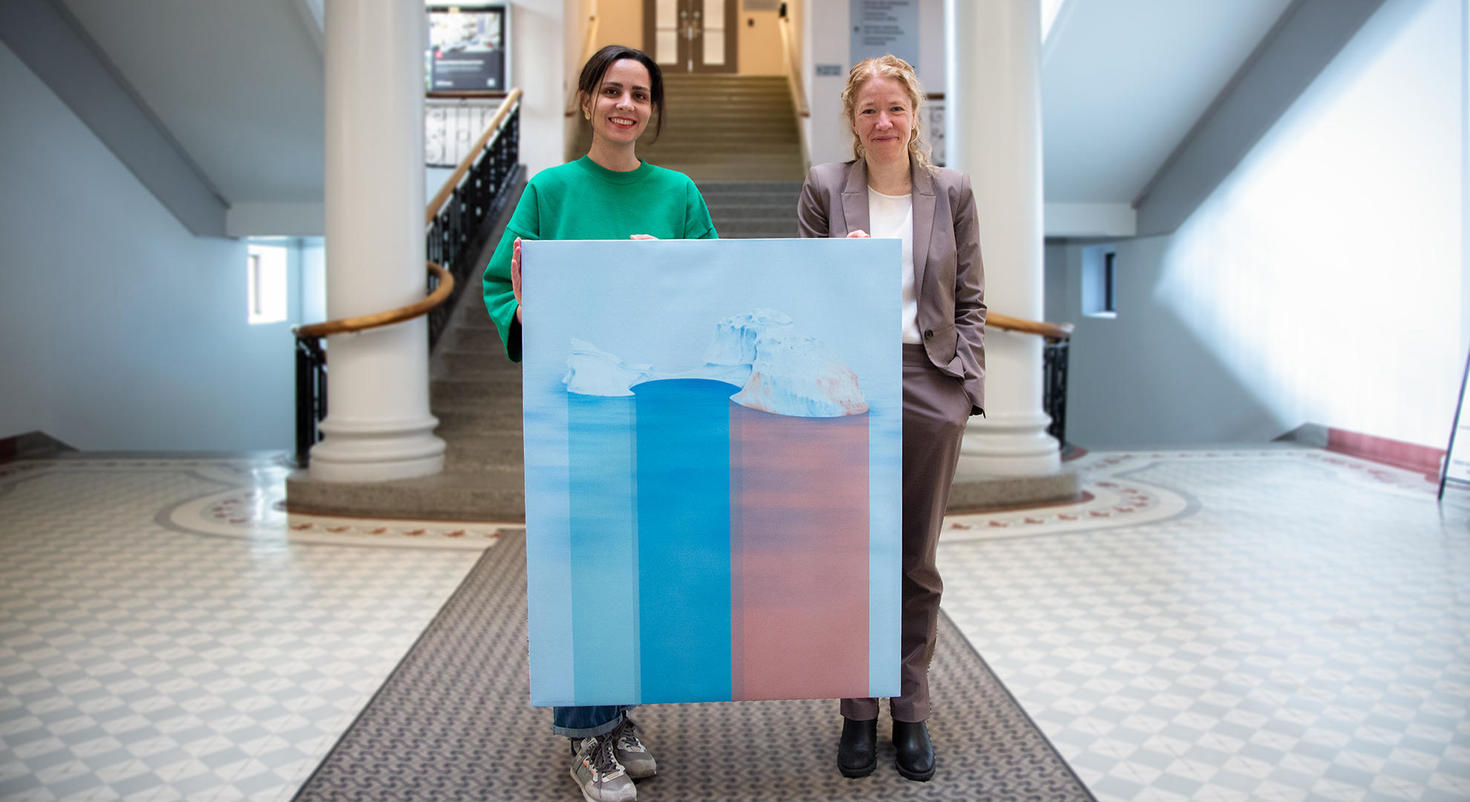 Mina Moshajjari, étudiante en arts visuels, et Julie St-Pierre, vice-présidente intérimaire à la recherche et de l'innovation, tenant l'œuvre gagnante de Mina, « Nature's response », dans le hall d'entrée de l’édifice Tabaret de l'Université d'Ottawa. L'œuvre représente un iceberg avec une vue au-dessus et au-dessous de l'eau, dans des tons roses et bleus.
