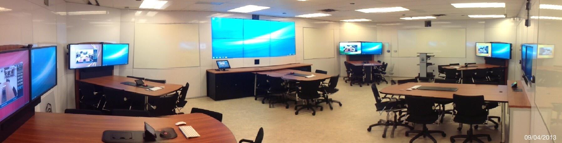 A modern classroom with multiple desks, computers, and wheeled office chairs, featuring large screens on the walls displaying various images