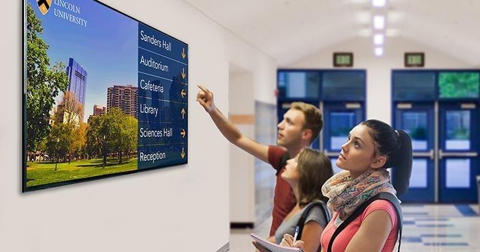 Three students standing in front of a digital information board inside a building