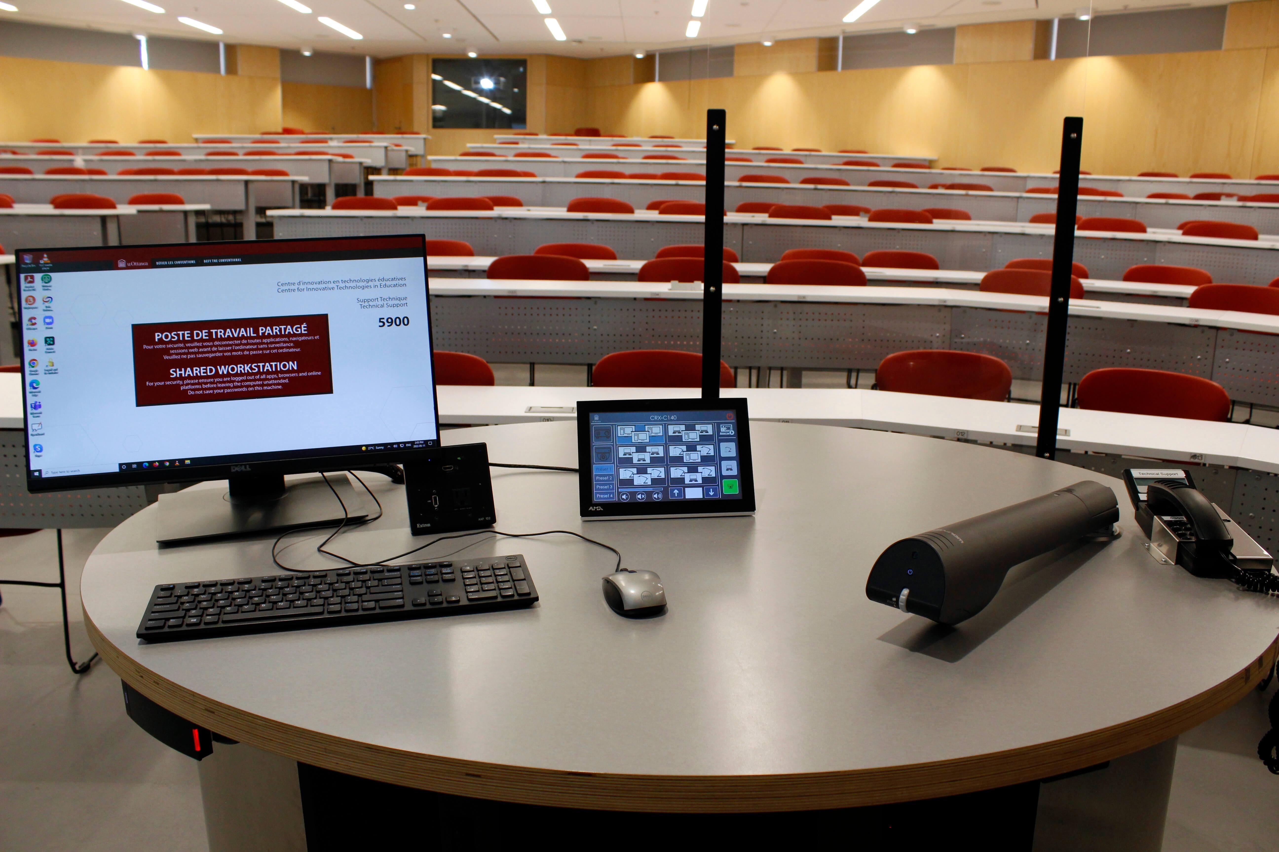 A lecture hall with red seats and a speaker’s podium equipped with a computer, microphone, and a projector screen