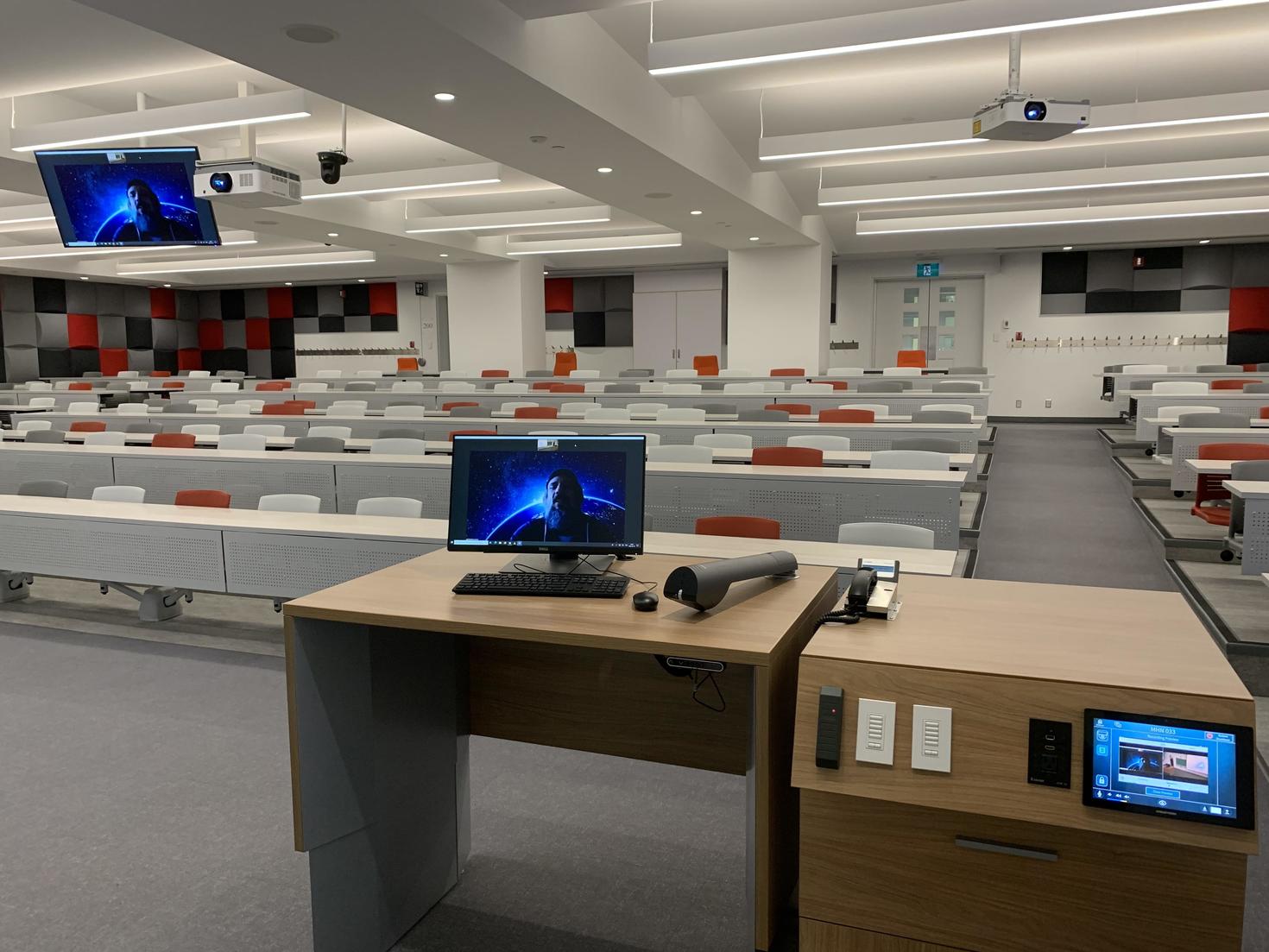 lecture hall with rows of empty white desks and red chairs, a podium with a computer monitor in the foreground, and projection screens displaying a blue graphic