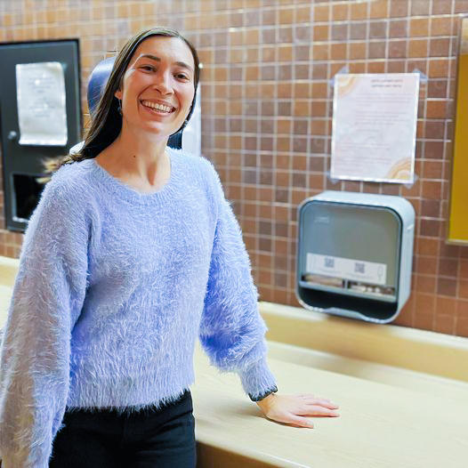 Woman smiles in front of a dispenser with free menstrual products in a washroom