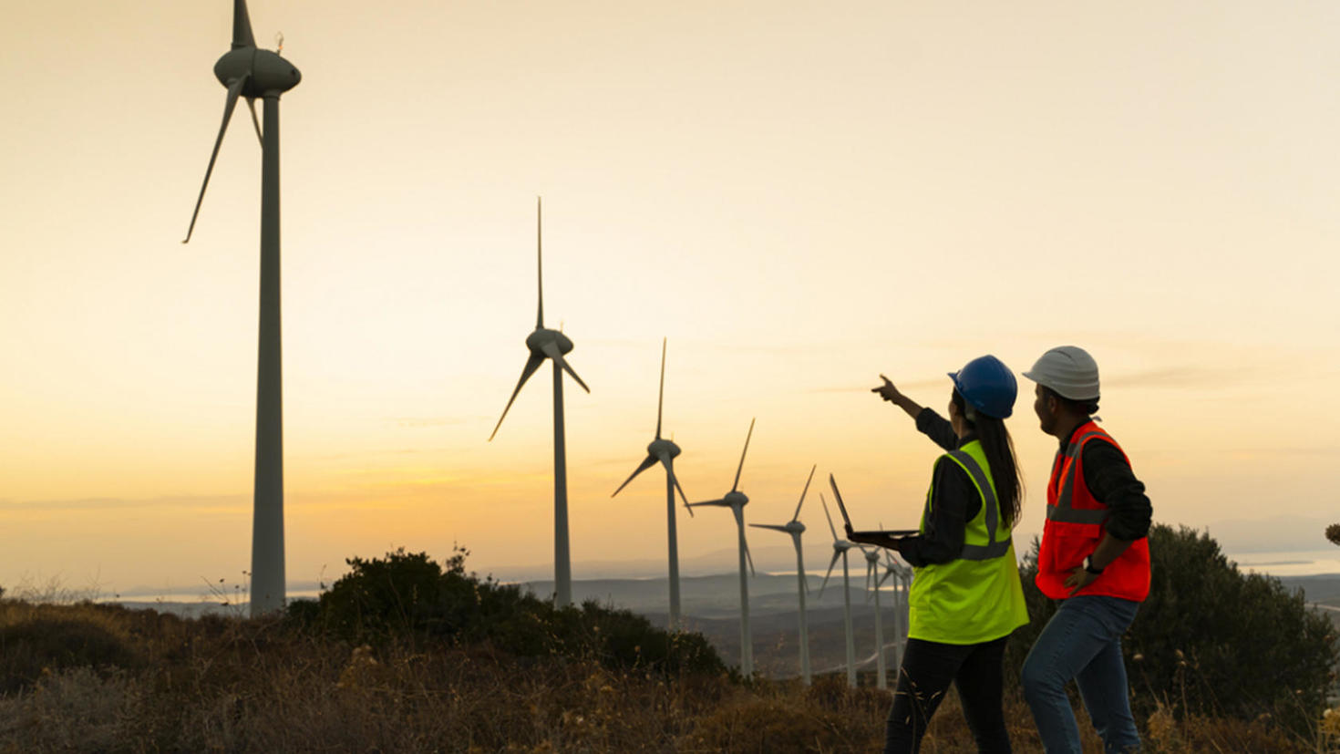 Two engineers in safety gear inspect wind turbines at sunset