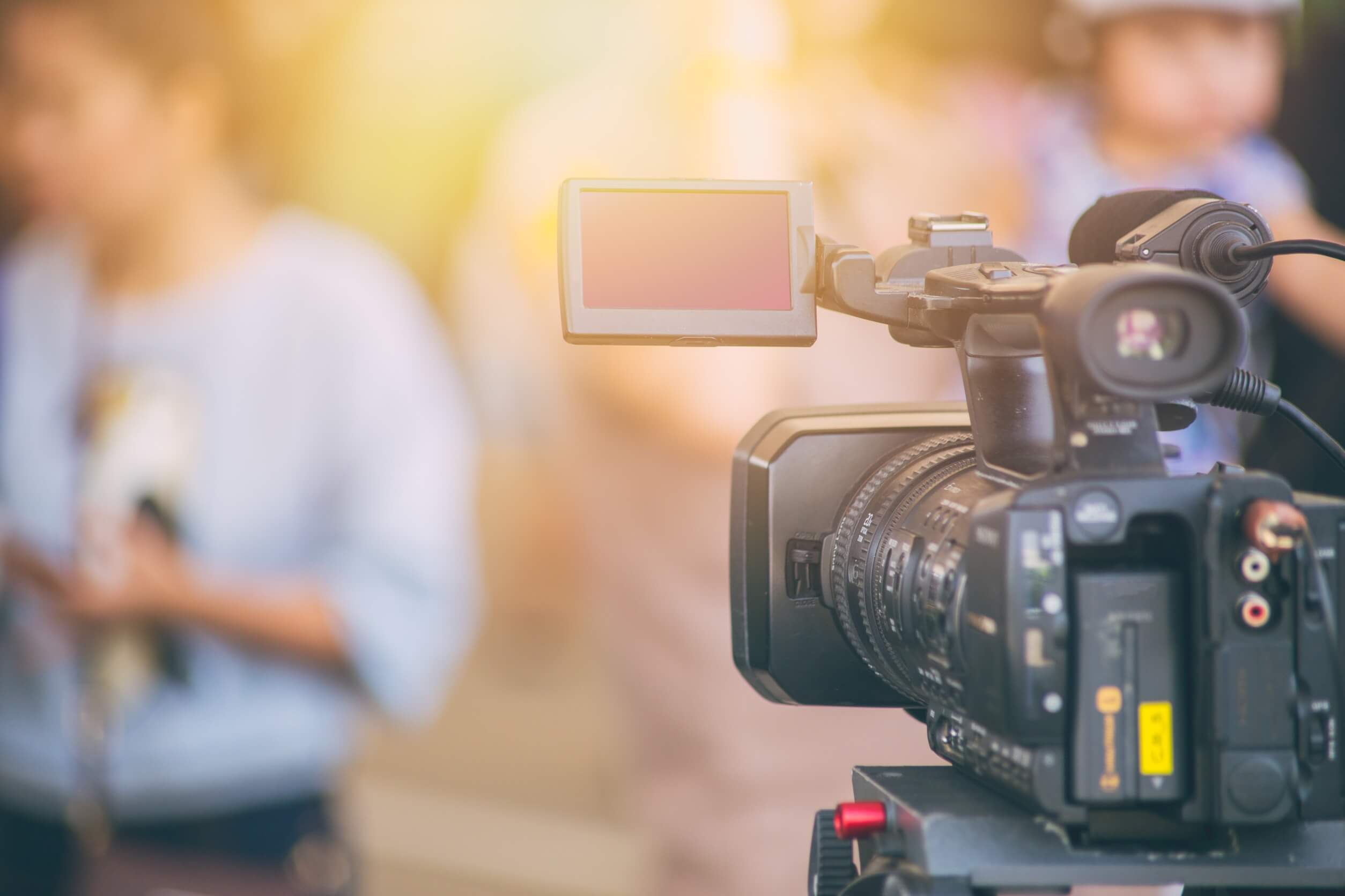 Une caméra vidéo professionnelle sur un trépied, mise au point au premier plan, avec un arrière-plan flou montrant une personne tenant un microphone.