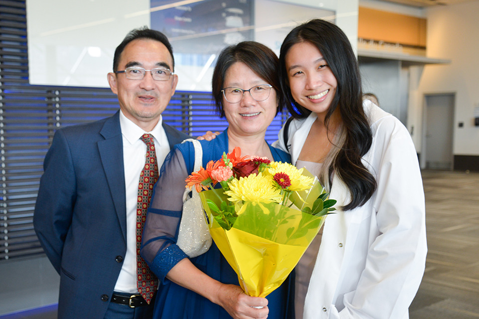 A medical learner in her white coat poses with her parents.