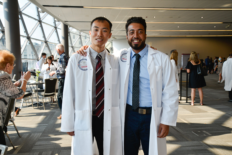 Two medical learners pose wearing their white coats.