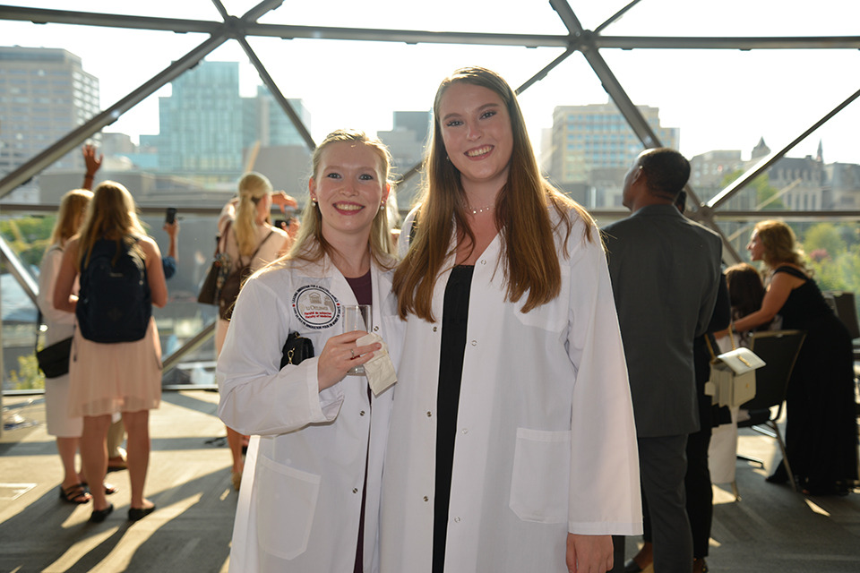 Two medical learners pose wearing their white coats.