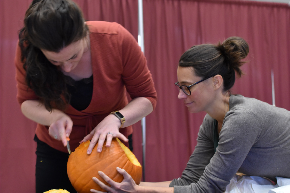 Cynthia Abraham leans over a table, holding a pumpkin, while Kayla Desnoyers cuts into the pumpkin with a carving knife.