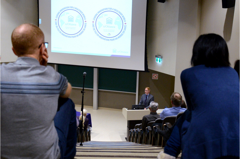 Two people sit, facing away from the camera, in the back row of a large auditorium. Between them, the figure of Dr. Bernard Jasmin can be seen at a podium at the front of the auditorium. A large screen at the front of the auditorium shows a projection of