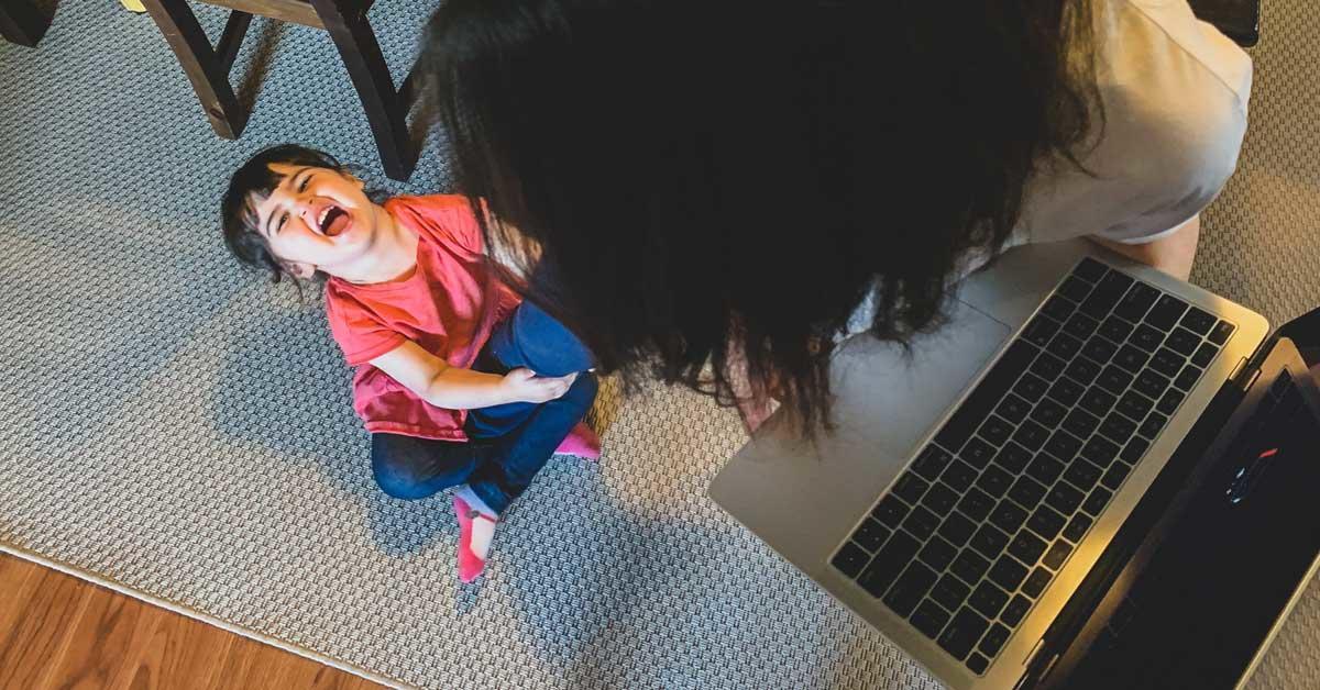 A young child sits on the floor while pulling on her mother's leg. The mother is trying to work on her laptop.