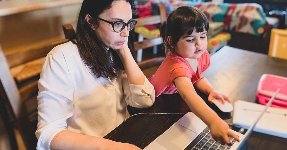 A woman working on her laptop as her young daughter sits beside her, holding the computer mouse and pointing to the laptop screen.