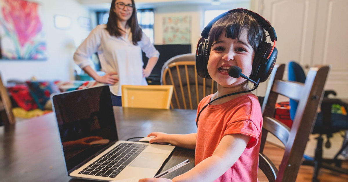 A young child wearing headphones sits in front of a laptop as her mother watches her.